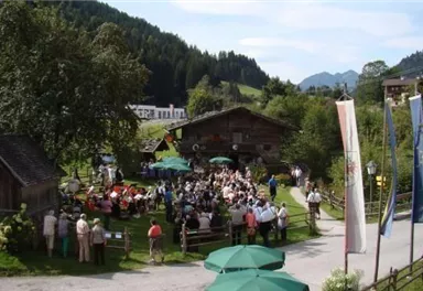 Eine große Menschenmenge feiert im Freien, umgeben von grünen Wiesen und Bergen. Es gibt Tische mit Sonnenschirmen und traditionelle Holzgebäude in der Nähe.