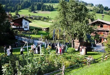 Eine festliche Veranstaltung im Freien mit Gästen in einem grünen Garten. Im Hintergrund sind traditionelle Bauernhäuser und sanfte Hügel zu sehen.