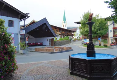 A beautiful village square with a fountain in the foreground. In the background, traditional buildings and a church tower can be seen.