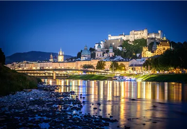 Eine malerische Stadtansicht von Salzburg bei Nacht. Die Lichter spiegeln sich im Fluss, während die Festung im Hintergrund leuchtet.
