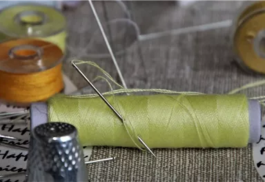 A sewing spool with green thread and a needle is lying on a table. In the background, there are more sewing spools in various colors.