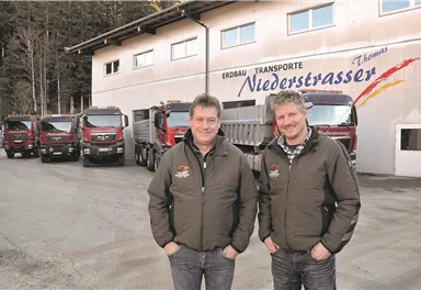 Zwei Männer stehen vor einem Lastwagenbetrieb mit mehreren Lkw im Hintergrund. Das Gebäude hat die Aufschrift „Niederstrasser Transporte“.