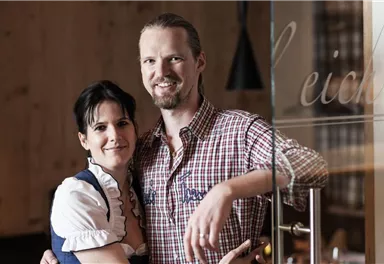 A smiling couple stands in front of a glass wall. The man is wearing a checkered shirt, the woman a traditional dress.