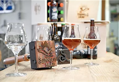 Ein Tisch mit verschiedenen Biergläsern und Flaschen. Im Vordergrund stehen zwei gefüllte Gläser mit Bernsteinfarbenem Bier.