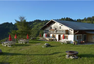 Eine gemütliche Berghütte umgeben von grünen Wiesen und Bäumen. Rote Schirme und Holztische laden zum Verweilen im Freien ein.