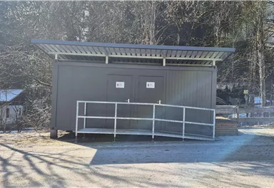 Eine moderne, graue Umkleidekabine steht auf einem befestigten Platz. Umgeben von Bäumen und einem klaren Himmel wirkt sie einladend und funktional.