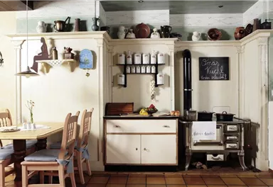 A cozy kitchen with a wooden table and chairs. On the wall, there are shelves with pots and herbs as well as a stove.
