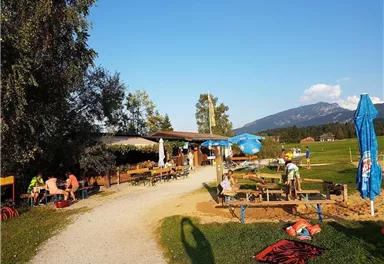 Ein gemütlicher Biergarten in der Natur mit Tischen und Bänken. Im Hintergrund erstreckt sich eine Wiese und die Berge sind sichtbar.
