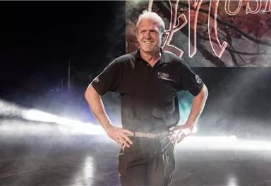 Ein Mann steht auf der Bühne und lächelt. Im Hintergrund sind Lichteffekte und ein großes Banner zu sehen.