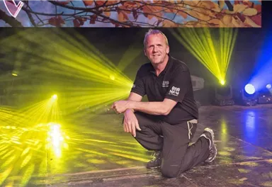 Ein lächelnder Mann kniet auf einer Bühne. Bunte Lichtstrahlen strahlen um ihn herum.