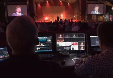 Eine Veranstaltung mit einem Musiker auf der Bühne. Im Vordergrund sind Techniker, die die Show mit Bildschirmen und Steuerungstechnik überwachen.