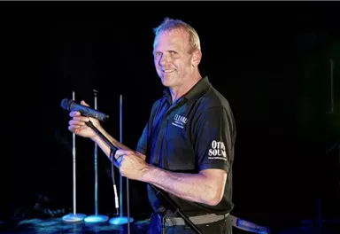 Ein Mann steht auf einer Bühne und hält ein Mikrofon in der Hand. Er lächelt und wirkt bereit für eine Performance.