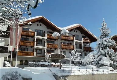 Ein hübsches Hotel im Schnee mit balkonartigen Fenstern und einer gemütlichen Holzkonstruktion. Umgeben von schneebedeckten Bäumen und einem klaren blauen Himmel.