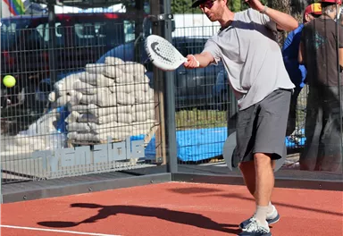 Ein Mann spielt Padel-Tennis auf einem roten Platz. Die Sonne scheint und im Hintergrund sind Bauarbeiten zu sehen.