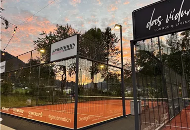 Ein Tennisplatz mit einem hellen, orangefarbenen Belag ist umgeben von einem Zaun. Im Hintergrund ist ein schöner Sonnenuntergang und einige Bäume zu sehen.