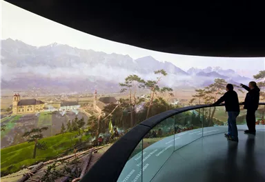 Zwei Personen stehen in einem modernen Ausstellungsraum und schauen auf eine beeindruckende Landschaft mit Bergen und einer kleinen Kirche. Die Aussicht ist lebendig und detailreich, was die Schönheit der Natur hervorhebt.