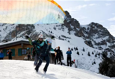 Eine Person startet mit dem Gleitschirm in einer schneebedeckten Berglandschaft. Im Hintergrund sind Berge und eine Hütte zu sehen, während andere Personen zuschauen.