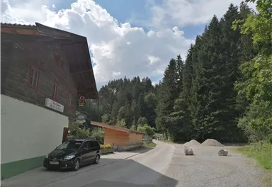 Eine kurvenreiche Straße führt durch eine malerische Landschaft mit dichten, grünen Wäldern. Auf der linken Seite steht ein rustikales Holzhaus.