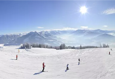 Eine wunderschöne Schneelandschaft mit Skifahrern und Bergen im Hintergrund. Der Himmel ist klar und sonnig.