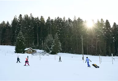Eine winterliche Landschaft mit Schnee und Bäumen im Hintergrund. Kinder spielen und ziehen einen Schlitten über die schneebedeckte Fläche.
