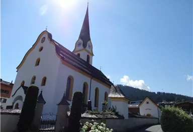 A picturesque church with a pointed tower in the foreground. The sky is blue and sunny, surrounded by green hills.