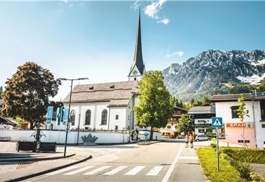 A picturesque church with a tall tower stands in the foreground. In the background, impressive mountains and a clear sky can be seen.