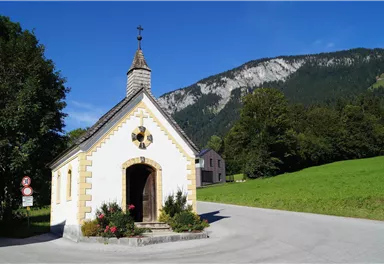 A small chapel stands at a street corner, surrounded by green meadows and trees. In the background, mountains are visible under a clear blue sky.