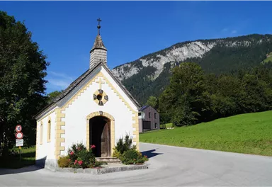 A small chapel stands by a road, surrounded by green meadows and trees. In the background, mountains and a blue sky can be seen.