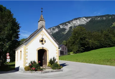 A small chapel stands at an intersection in nature. In the background, green meadows and mountains can be seen.