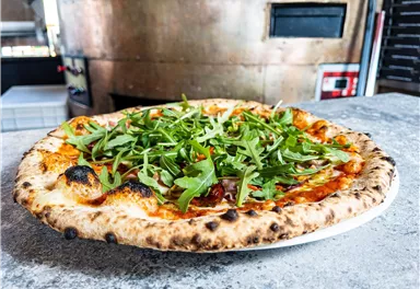 A freshly baked pizza with arugula on a plate. In the background, a pizza oven is visible.