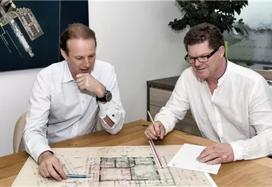 Two men are sitting at a table, looking at an architectural plan. They are discussing and taking notes on paper.