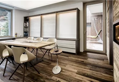 A modern dining room with a large table and white chairs. The glass windows provide a view outside to a terrace.