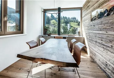 A modernly designed dining area with a wooden table and brown chairs. Large windows offer a view of the surrounding mountains and nature.