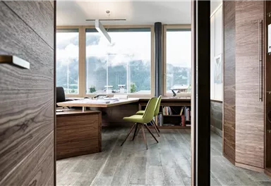 A modern office with large windows and a view of the mountains. The space is designed with wood and includes a desk as well as a green chair.