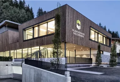 A modern building with a wooden façade and large windows. It is surrounded by trees and looks inviting.