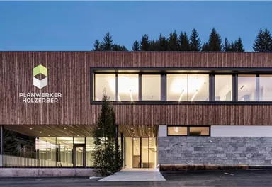 A modern building with wood cladding and large windows. In the foreground, there is a green plant and trees are visible in the background.