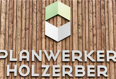 A wooden sign with the inscription "PLANWERKER HOLZERBERG". Above it is a geometric logo in green and white.