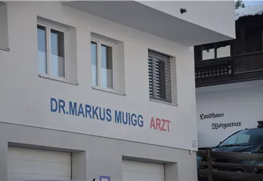The image shows the exterior view of a doctor's office with the inscription "Dr. Markus Muigg Arzt". The facade is designed in a simple and modern style.