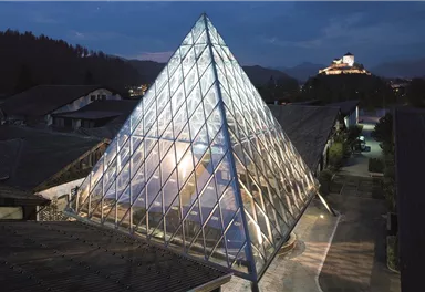 Ein modernes, pyramidenförmiges Gebäude aus Glas steht im Vordergrund. Im Hintergrund ist eine Burg auf einem Hügel zu sehen, während der Himmel in Dämmerungshfarben leuchtet.