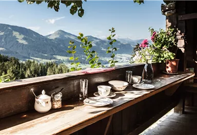 Ein schöner Tisch mit Geschirr und Gläsern auf einer Holzterrasse. Im Hintergrund sind Berge und eine klare, sonnige Aussicht zu sehen.