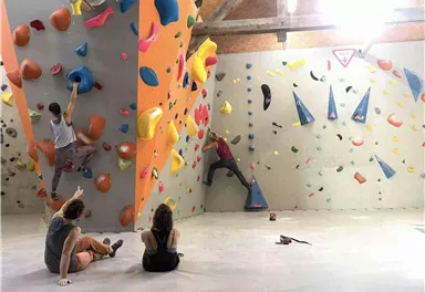 A modern bouldering center with two climbing walls. Two climbers are active while two spectators are sitting back relaxingly.