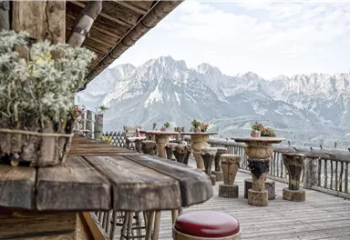 A wooden terrace with rustic tables and flowers. In the background, impressive mountains rise under a clear sky.