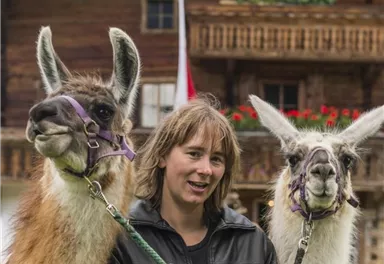A woman stands with two llamas in front of a wooden house. The background is decorated with flowers.