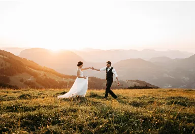 A bridal couple dances happily on a green meadow in the mountains. In the background, the setting sun glows.