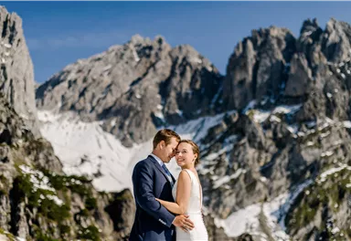 A bride and groom pose in front of an impressive mountain landscape. The peaks are snow-covered and the sky is clear.