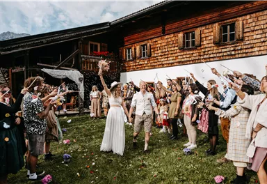 A bride and groom celebrate in front of a traditional wooden house. Surrounded by cheerful guests who cheer with flowers and confetti.