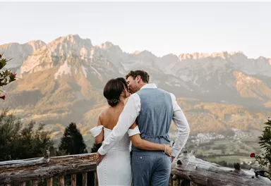 A bride and groom are standing on a porch and embracing each other. In the background, majestic mountains and a picturesque landscape can be seen.