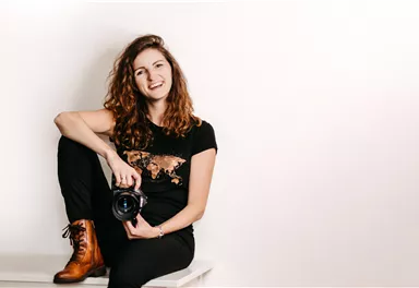 A smiling woman with curly hair is sitting on a table and holding a camera. She is wearing a black T-shirt and brown boots.
