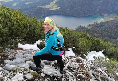 Eine aktive Person wandert einen steilen Berg hinauf. Im Hintergrund sieht man einen schönen See und bewaldete Berge.