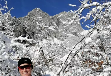 Eine Person steht in einem schneebedeckten Wald mit Bergen im Hintergrund. Der Himmel ist klar und blau.